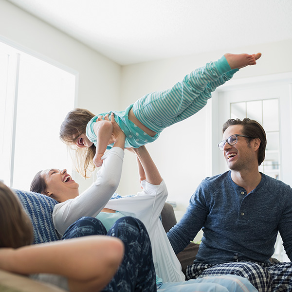 A woman sitting on a couch joyfully lifting a young child in the air as other family members look on