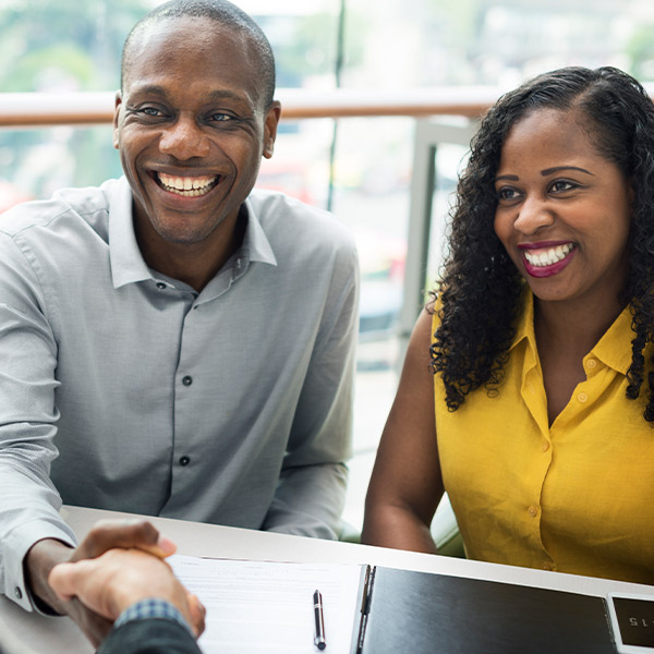 Smiling couple seated at desk, man shakes hand with man behind the desk