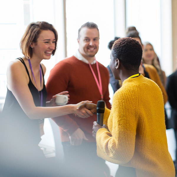 woman in yellow sweater holding microphone shaking hands with a woman at a conference